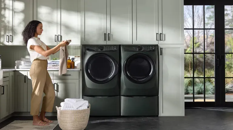 Electrolux ELFW7738AA Front-Load Washer and matching dryer in a black steel finish, installed as a set on pedestals in a bright, modern laundry room with sage green cabinetry